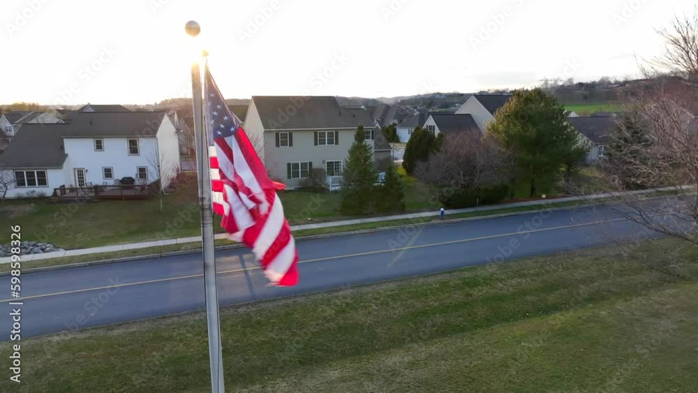 American flag waving in front of neighborhood in USA during sunset. Beautiful establishing shot of housing in the United States. Aerial orbit shot. American flag waving in front of neighborhood in USA during sunset. Beautiful establishing shot of housing in the United States. Aerial orbit shot.