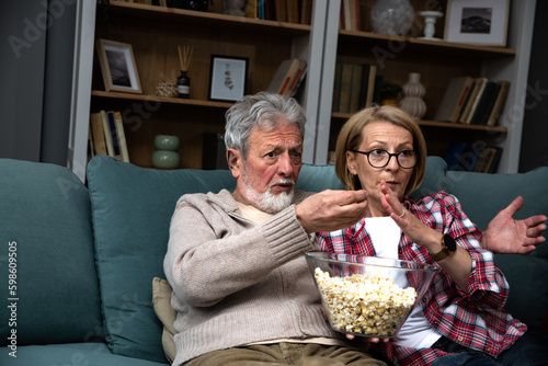 Mature married couple entertaining observing terrified TV program. Senior husband and wife watching horror movie or film at home eating popcorn and making scary facial expressions