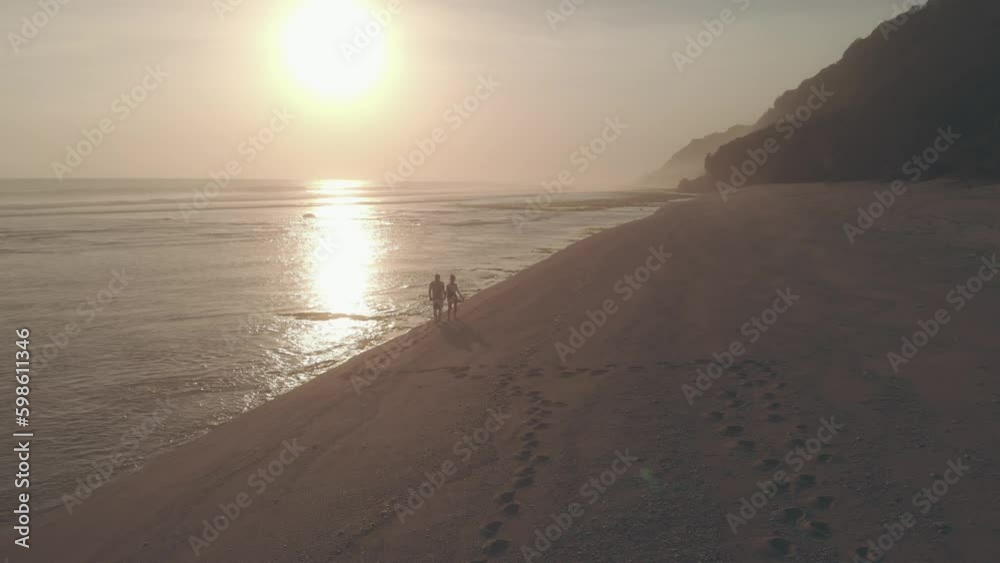 Drone aerial view. Silhouettes of couple in love walking on the beach at sunset. Man and woman holding hands walk, leaving footprints on sand. Sun reflecting in the sea. Concept of romance and love Drone aerial view. Silhouettes of couple in love walking on the beach at sunset. Man and woman holding hands walk, leaving footprints on sand. Sun reflecting in the sea. Concept of romance and love