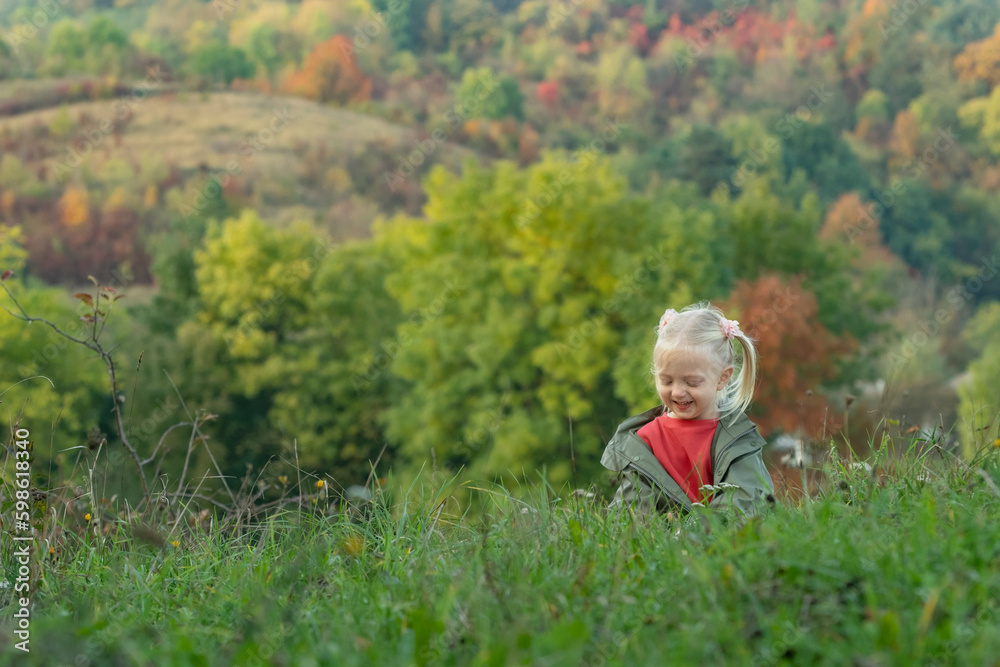 Little blonde girl plays on grass in clearing on warm autumn day. Child spends time in nature.Landscape of autumn forest Little blonde girl plays on grass in clearing on warm autumn day. Child spends time in nature.Landscape of autumn forest