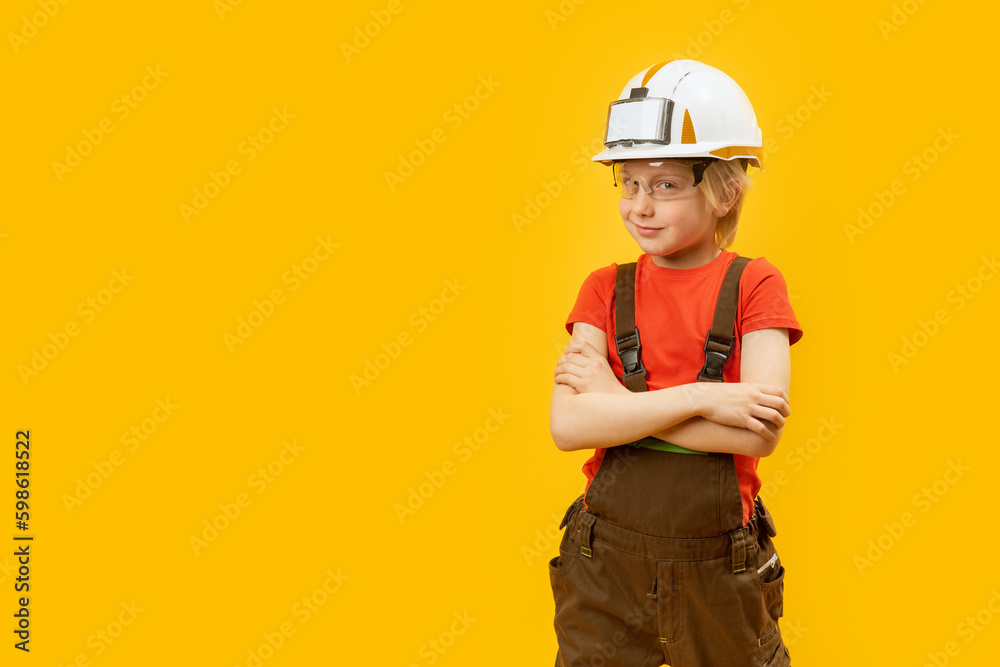 Portrait of smiling boy in protective helmet, glasses and jumpsuit like worker. Yellow background. Copy space, mock up Portrait of smiling boy in protective helmet, glasses and jumpsuit like worker. Yellow background. Copy space, mock up