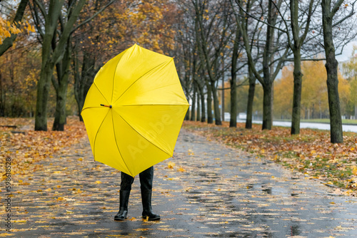 Child hid behind large yellow umbrella on autumn park and fallen leaves background. Back view