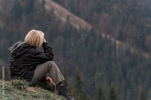 Blond boy sits at hillside and looks at the distance. Side view on boy in mountains. Vacation with children
