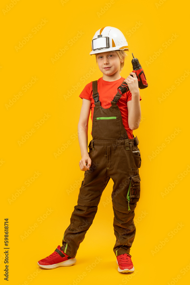 Portrait of teenage boy in work jumpsuit and protective helmet holds drill. Child is like worker builder on yellow background.