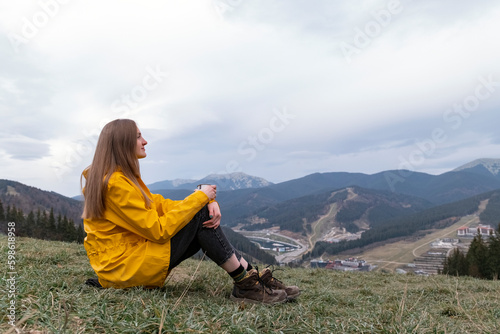 Tourist young woman sits on hillside with cup in autumn. Female traveler looks at the mountains.
