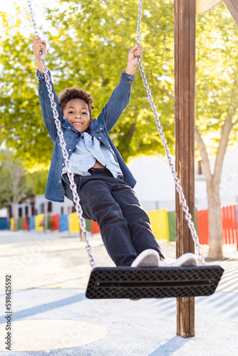 A happy, dark-skinned boy with curly afro hair between the ages of 5 and 6 is standing on a swing in an outdoor public park. Concept of extracurricular activities.