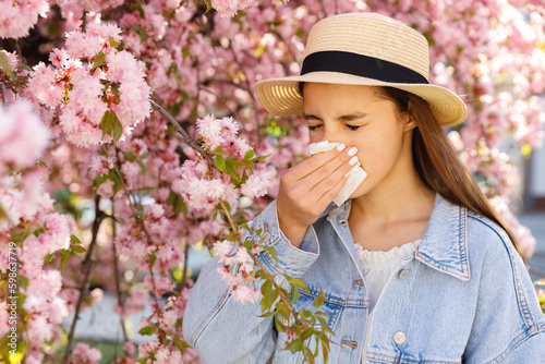 Symptoms from Seasonal Allergy. Sick Girl Sneezing in Napkin while Standing near Blooming Sakura Tree on City Street