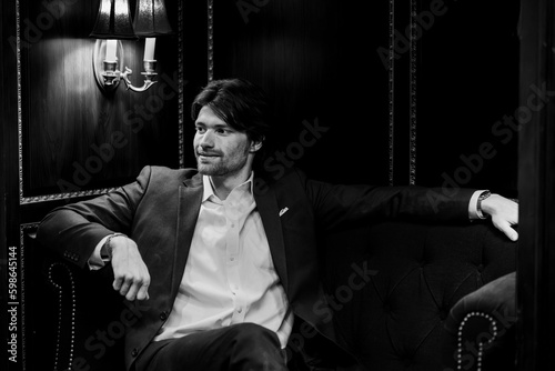 Black and White Man in Suit Business Casual Casually Looking Off with a Light Grin Smile Happy Satisfied in Library with Vintage Antique Dark Light Fixture Moody Feel Jacket Relaxed Professional