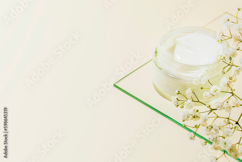 Cosmetic glass jar with natural cream on glass shelf on light beige background with white flowers. Organic beauty product and skin friendly cosmetics concept. Mockup, copy space, close up