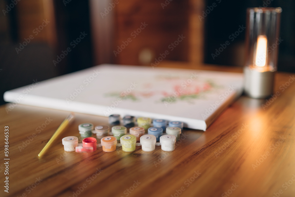 Fototapeta premium Painting by numbers. A set for painting a picture, including a canvas, paints of different colors, brushes. Side view. Lamp in the background.
