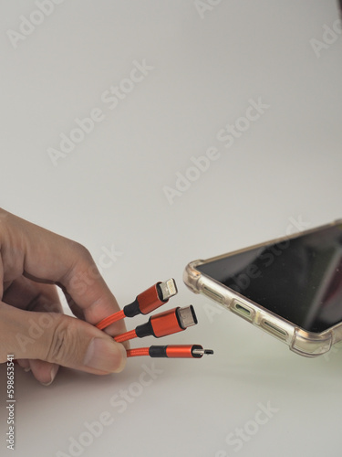 A person is ready to charge a mobile phone on a white background. Close up a cable with 3 connectors: Lightning / USB-C / Micro USB, will be plugged into a smartphone.