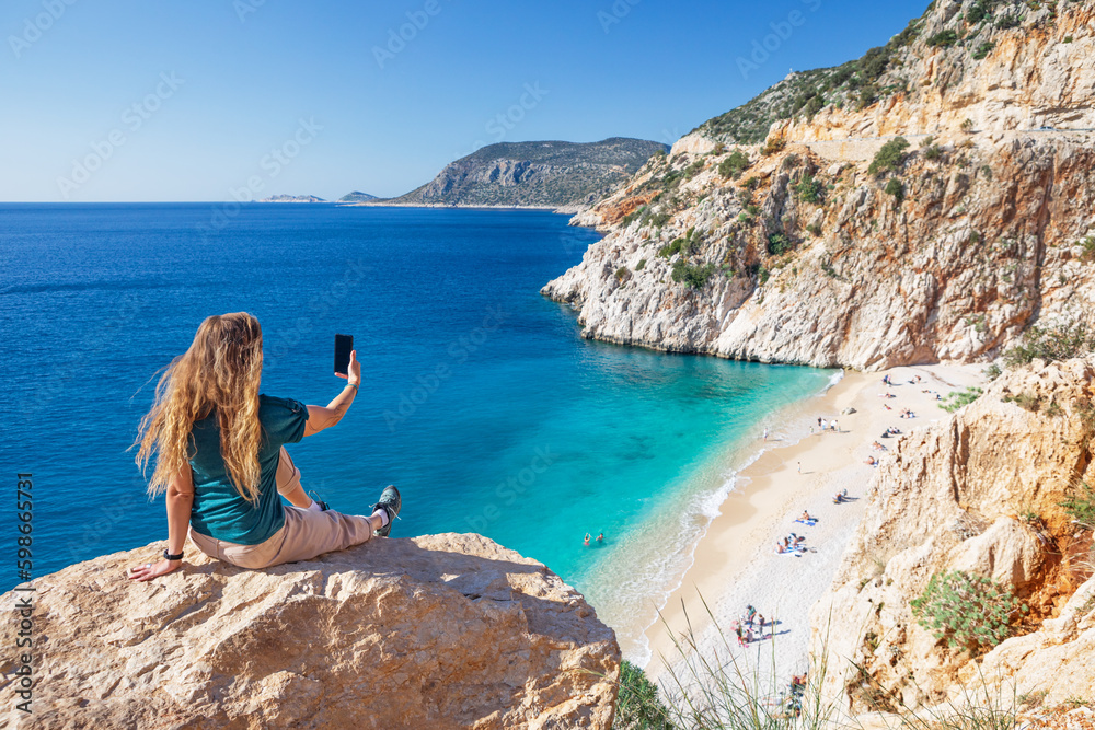 Naklejka premium Young woman making selfie photo by smartphone over Kaputas beach, Lycia coast. Summer day walk by Lycian way at family vacation in Mediterranean Sea, Kas, Antalya region, Turkey