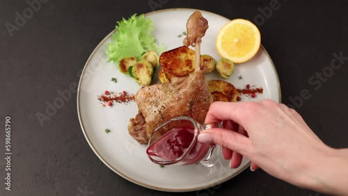 Duck leg confit with Brussels sprouts, baked potatoes, thyme, orange and lingonberry sauce. Traditional French cuisine. Woman pours sauce on a plate. Selective focus, close-up, black background.