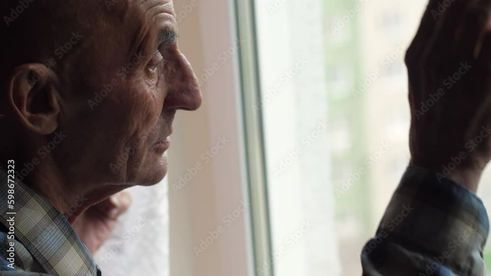 the pensioner looks out the window and waves his hand while inside. an elderly person is waiting for close people, relatives, real pensioners, a visit to the parents of the elderly