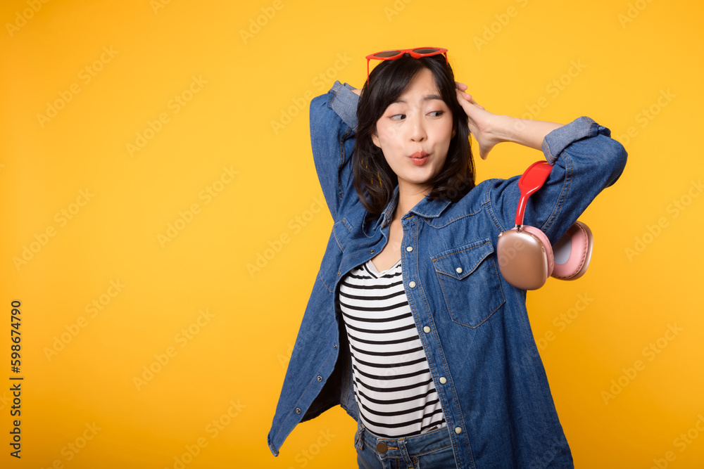Fashion portrait young asian woman wearing denim clothes, red glasses and headphone isolated on yellow background. Lifestyle people concept.