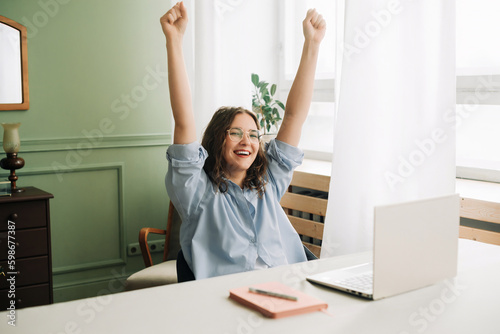 Joyful young woman celebrating success while using laptop computer for remote work or study. celebrating reaching a goal, winning online, getting good news by email, hands up, feeling euphoric.