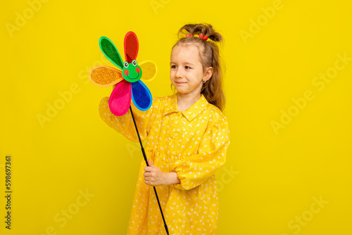 Photo of little cheerful girl in a beautiful yellow summer dress happy positive smile hold paper toy windmill on white background. Summer fashion for children.