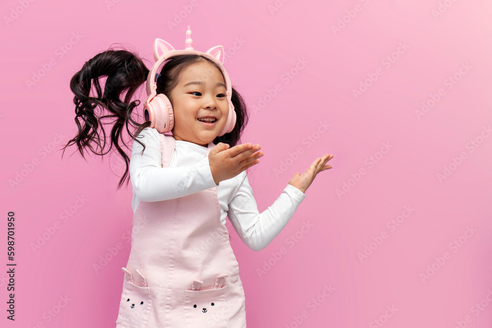 asian girl in pink children's headphones and glasses listens to music and dances on pink isolated background - obrazy, fototapety, plakaty 