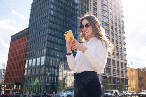 girl in glasses and white shirt is standing on the street in the city and using smartphone, woman is typing message