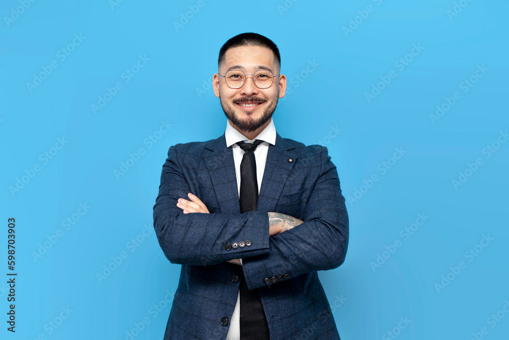 successful asian businessman in suit and glasses stands with his arms crossed on blue isolated background successful asian businessman in suit and glasses stands with his arms crossed on blue isolated background