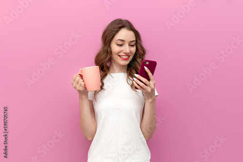 young cute woman in white t-shirt holds cup with drink and uses smartphone on pink isolated background