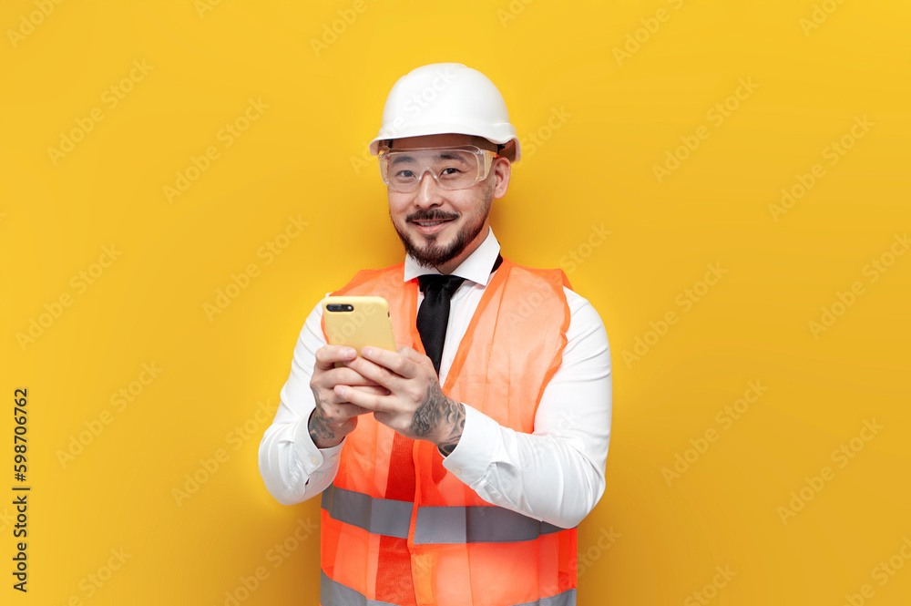 asian foreman in uniform uses smartphone on yellow isolated background, korean civil engineer in hard hat asian foreman in uniform uses smartphone on yellow isolated background, korean civil engineer in hard hat