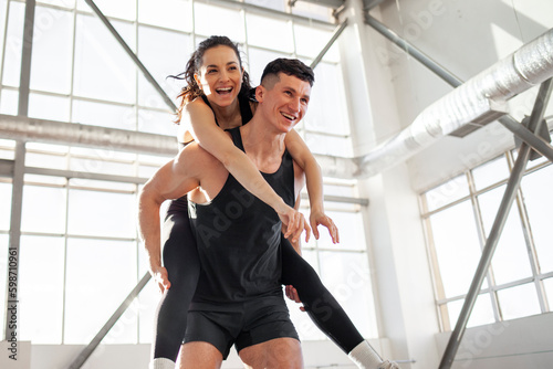 happy athletic couple in sportswear runs and rejoices in bright gym, the guy carries the girl on his back and smiles © Богдан Маліцький