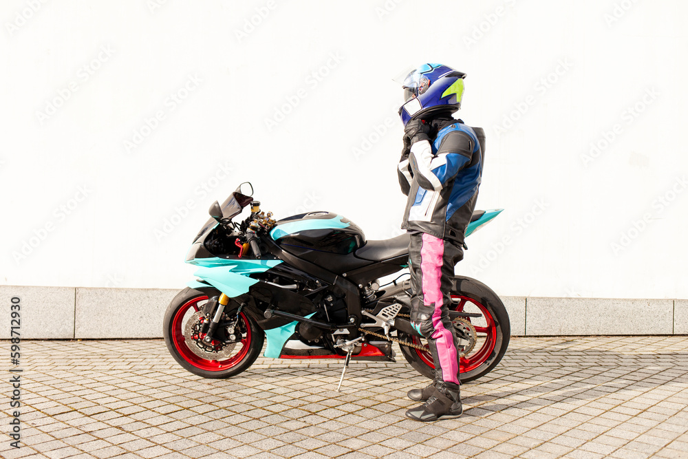 guy motorcyclist in professional protective equipment and helmet stands near sports motorcycle against white wall