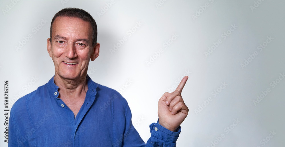 Happy and smiling mature adult caucasian male with blue shirt points away with finger over white grey background with copy space Happy and smiling mature adult caucasian male with blue shirt points away with finger over white grey background with copy space