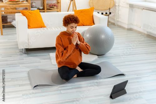 Yoga mindfulness meditation. Young healthy african girl practicing yoga at home. Woman sitting in lotus pose on yoga mat eyes close hands in prayer meditating indoor. Girl doing breathing practice
