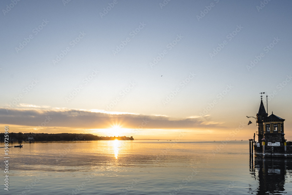 Panoramic sunrise view from Imperia statue to lighthouse at harbor entrance and Lake Constance in early morning hours. Steamer harbor, Constance, Baden-Württemberg, Germany, Europe. Panoramic sunrise view from Imperia statue to lighthouse at harbor entrance and Lake Constance in early morning hours. Steamer harbor, Constance, Baden-Württemberg, Germany, Europe.