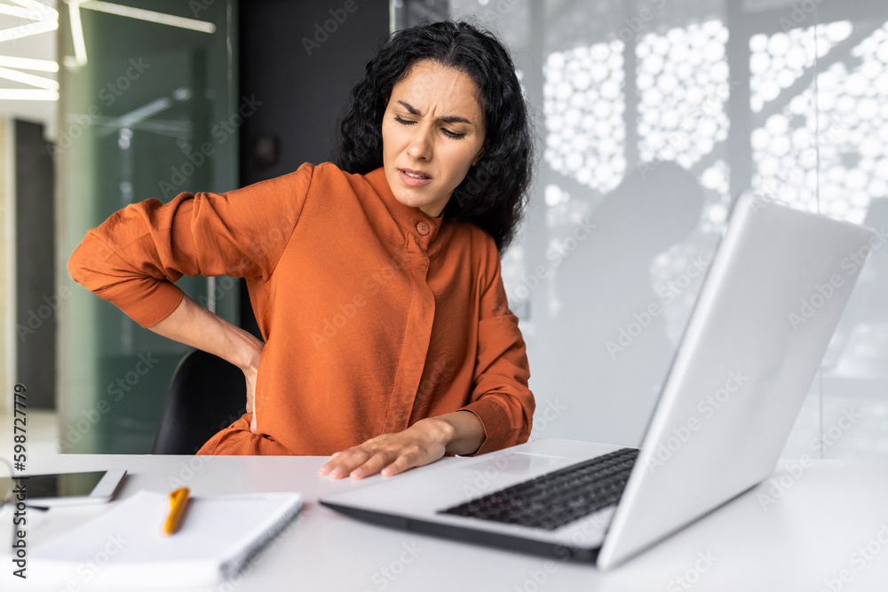 Overtired hispanic woman working late, business woman having severe back pain, female worker inside office sitting at table using laptop at work Overtired hispanic woman working late, business woman having severe back pain, female worker inside office sitting at table using laptop at work