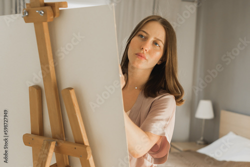 Young close-up woman artist painting on canvas on the easel at home in bedroom - art and creativity concept