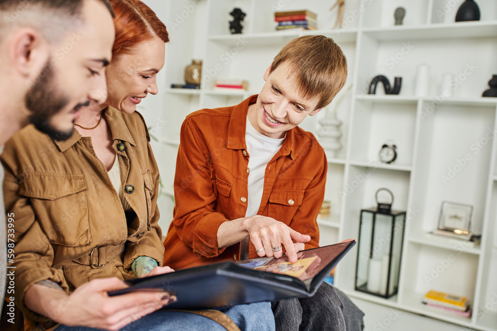 Obraz premium smiling gay man pointing at photo in album near happy mother and blurred boyfriend at home. 