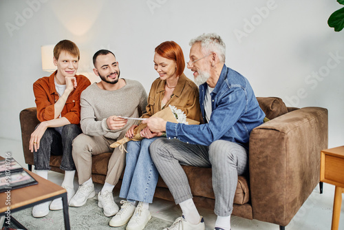 cheerful gay man giving wedding invitation to parents while sitting on couch in living room.