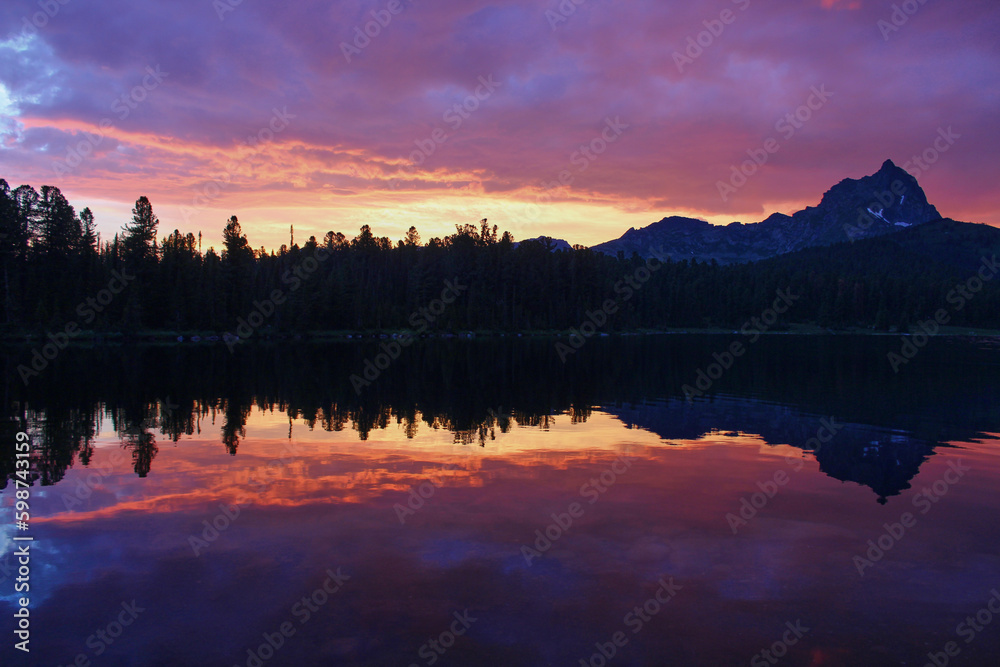 Incredibly beautiful sunset on a mountain lake. Trees and a rocky peak are reflected in the water. Purple sky
