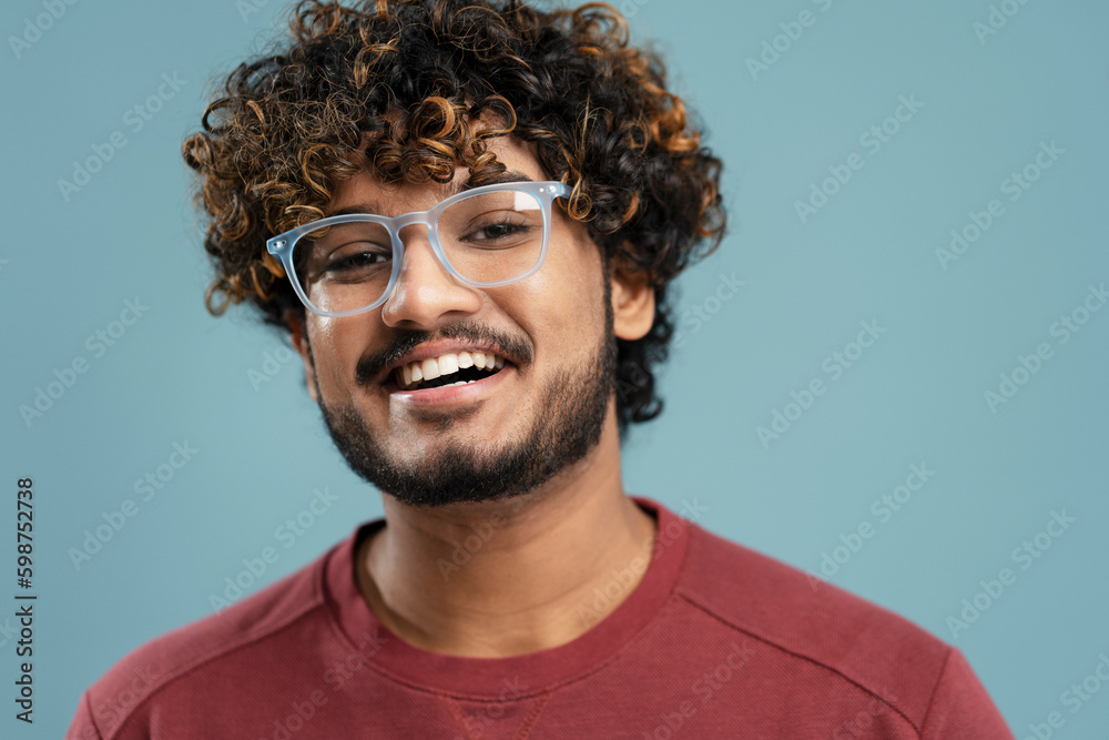 Obraz premium Portrait of handsome smiling curly haired Indian man, smart student wearing stylish eyeglasses looking at camera isolated on blue background. Vision concept