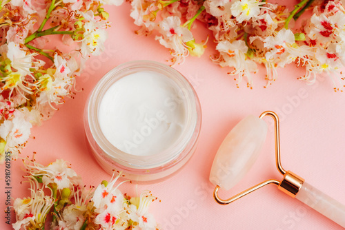 White cream jar and rose quartz facial roller with Horse chestnut flowers on pink background. Natural organic cosmetics concept. Top view, flat lay photo