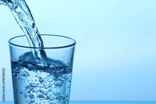 Pouring water into glass on light blue background, closeup. Space for text