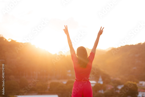 Happy woman with arms raised  making the victory sign with her fingers and sunset in the background. Concept of person overcoming problems and achieving success.