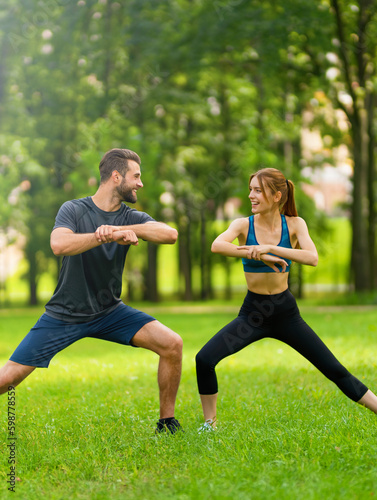 Image of young happy smiling couple, woman training with man or bearded coach trainer, doing squat fit exercise together, look at each other, outdoors. Fitness, sport, healthy lifestyle concept.