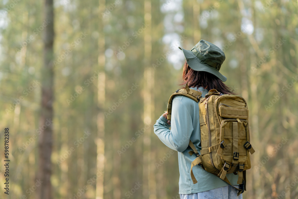 Naklejka premium Young Asian girl with a backpack and hat hiking in the mountains during the summer season, a traveler walking in the forest. Travel, adventure, and journey concept.