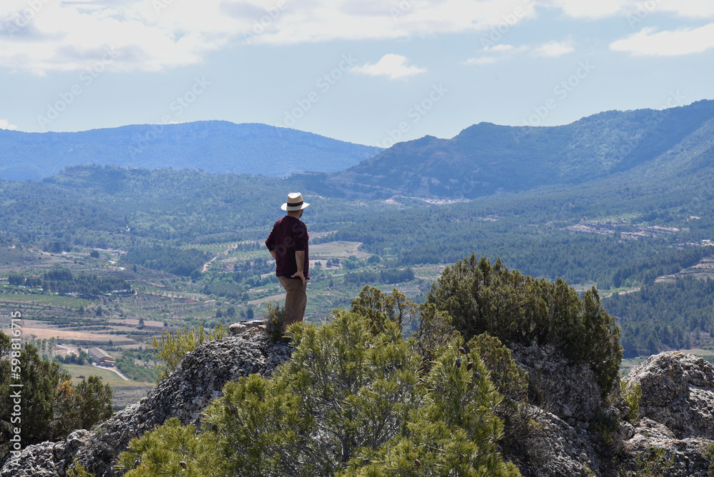 unrecognisable man in an explorer's cap, looking at the natural landscape on top of a small hill in a high place with a lot of countryside and forest.