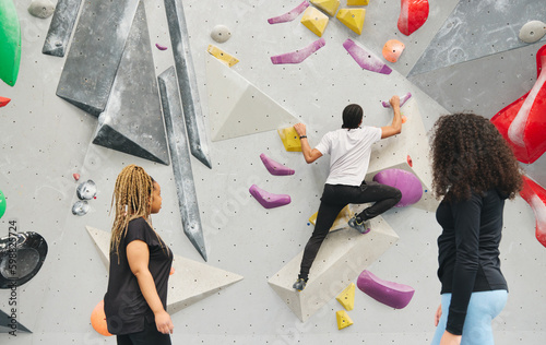 Multi-Cultural Group Watching Friend On Climbing Wall In Indoor Activity Centre