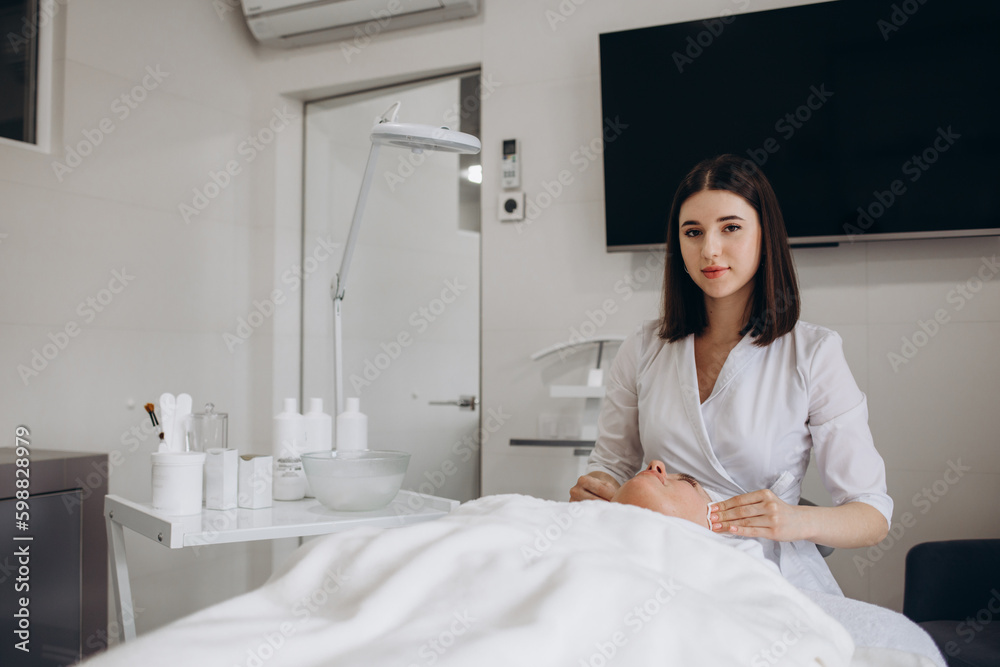 young woman cosmetologist in cosmetology clinic stands near the couch and smiles, girl dermatologist in doctor's uniform at the workplace.