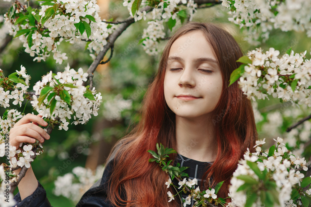 Fototapeta premium Young beautiful smiling girl standing with closed eyes among blooming white flowers and dreaming in spring park or garden, holding a branch with cherry blossom, tenderness and romance of springtime