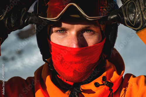 Fototapeta Naklejka Na Ścianę i Meble -  Portrait of a man with yellow eyes. Photo of a young serious sportsman in a helmet and ski goggles in his hands, against the backdrop of a snowy hill