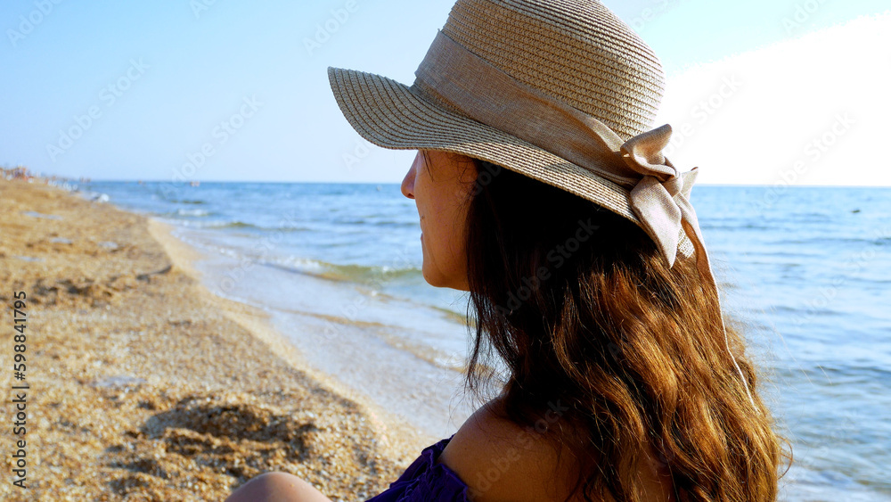 A girl with long brown hair in a hat sits on a deserted shore on the ...