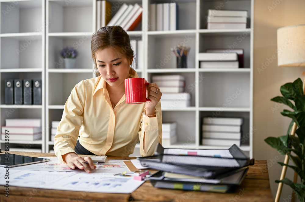 Focused Asian female accountant sipping coffee and analyzing business financial data on reports Focused Asian female accountant sipping coffee and analyzing business financial data on reports