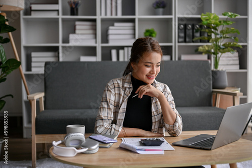 Happy Asian woman sits in her living room calculating taxes and managing household expenses.